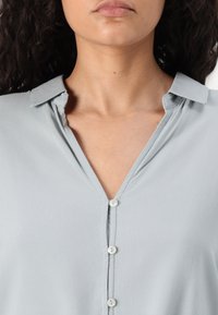 Light grey blouse with a V-neck collar, button closure, and soft fabric texture. Features a clean, simple design with no patterns.