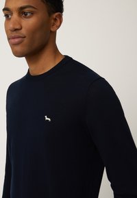 Marineblauer gestrickter Pullover mit Rundhalsausschnitt, der ein kleines weißes Emblem eines Hundes auf der linken Brust trägt. Glatte Textur, lange Ärmel.