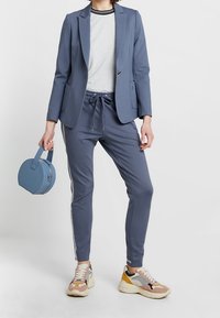 Jasnoniebieski żakiet na szarym podkoszulku, zestawiony z pasującymi joggerami z bocznymi paskami, akcentowany niebieską okrągłą torebką. Obuwie z wielokolorowymi panelami.