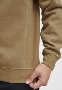 Brown sweatshirt with ribbed cuffs and hem. Smooth texture with visible stitching and a fitted design. Hand gripping the hem.