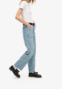 Jeans de mezclilla de corte amplio en azul claro con bolsillos frontales, un bolsillo trasero de parche y detalles de costura, combinados con zapatos negros y una camiseta blanca.