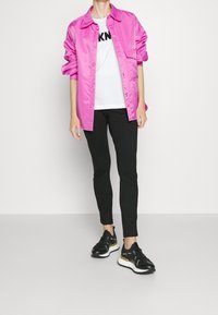 Różowa satynowa koszula zapinana na guziki, biała koszulka z czarnymi literami, czarne slim-fit spodnie oraz czarne sneakersy z białymi podeszwami i akcentami.