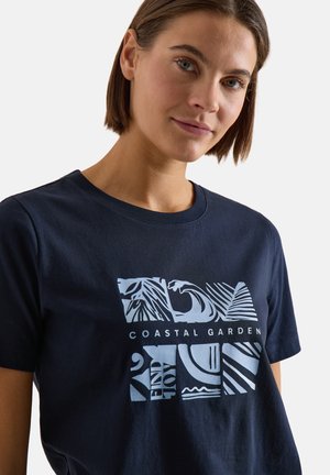 Jeune femme aux cheveux bruns courts portant un t-shirt bleu marine avec un motif côtier bleu clair et le texte « Coastal Garden ».