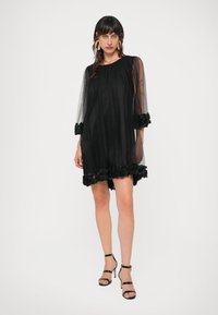 Μαύρο φόρεμα από τούλι με διαφάνειες και μακριά μανίκια, διακοσμημένο με λουλουδάτη δαντέλα στο τελείωμα και στους καρπούς, με άνετη εφαρμογή. Συνδυάζεται με μαύρες στιλέτο γόβες.