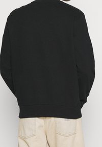 Felpa nera con colletto tondo, maniche lunghe e polsini a coste. Tessuto morbido e vestibilità rilassata. Indossata con pantaloni di colore chiaro.