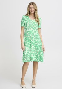 Vihreä kukallinen mekko, jossa on lyhyet hihat, V-pääntie ja liukuvakuvioinen helma. Valmistettu pehmeästä kankaasta, jossa on tekstuuri. Yhdistetty vaaleisiin kenkiin.
