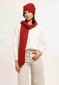 Red knit beanie and matching scarf displayed over a white sweater, featuring a textured fabric. Model wears light beige wide-leg trousers.