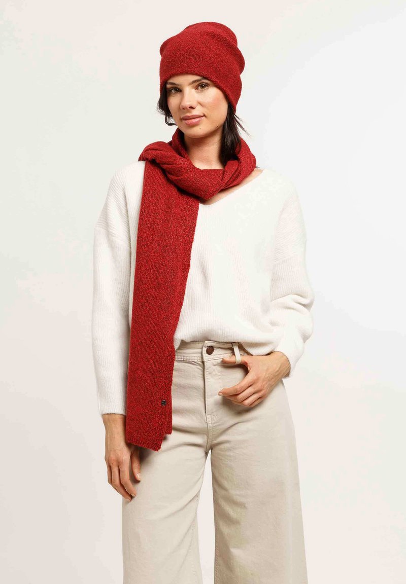 Red knit beanie and matching scarf displayed over a white sweater, featuring a textured fabric. Model wears light beige wide-leg trousers.