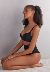 Zwarte naadloze bh en hoge taille slip van een gladde stof. Voorzien van verstelbare bandjes en een minimalistisch ontwerp. Model posed in zijprofiel.