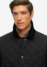 Czarna pikowana kurtka z kołnierzem z kordurowego materiału, z złotymi dodatkami i koszulą w kratę pod spodem.