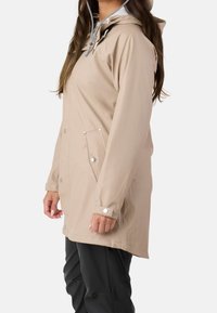 Femme portant un imperméable beige à capuche avec poches et poignets à boutons-pression, associée à un pantalon noir, debout de profil sur fond blanc.