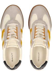 Scarpe sportive bianche e grigie con strisce gialle. Presentano materiali in suede e tela, design con lacci e un cappuccio sul tallone nero.
