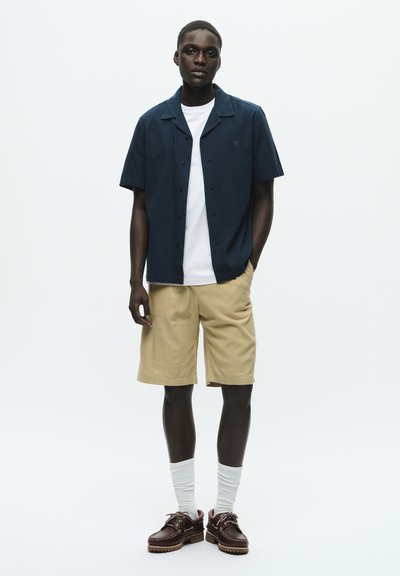 Jeune homme debout portant une chemise bleu marine à manches courtes, un t-shirt blanc, un short beige, des chaussettes blanches et des chaussures à lacets marron foncé.