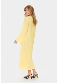 Long yellow dress with a pleated texture, featuring long sleeves and a flowing silhouette. Worn with black heeled shoes.