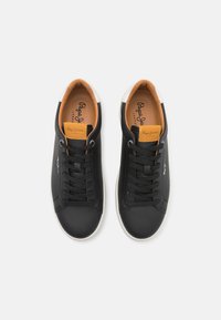 Fekete bőrsneaker narancssárga részletekkel, fehér gumitalppal, kerek orral és fekete fűzőkkel. Az oldalon és a nyelvén a márka logója található.
