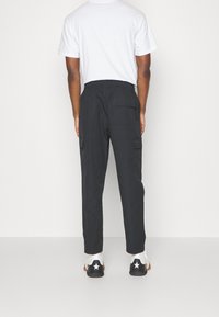 Man som bär svarta cargo-byxor, vit t-shirt och vita sneakers med svart stjärndetalj, stående mot en plain vit bakgrund.