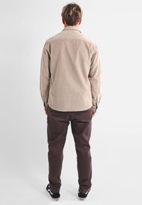 Camisa de pana beige con cuello abotonado, mangas largas y dobladillo recto; combinada con pantalones marrón oscuro y zapatillas negras.