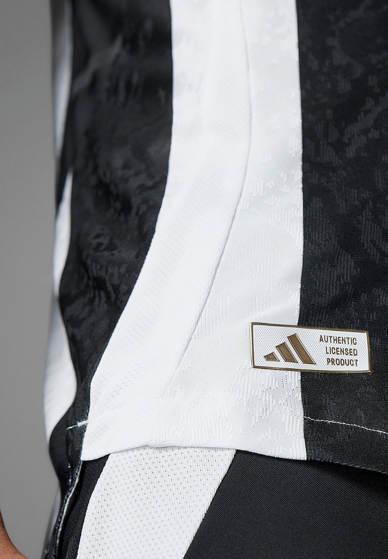 Sort og hvid atletisk skjorte med struktureret sort stof, glatte hvide detaljer og et guld "AUTENTISK LICENSERET PRODUKT" mærkat.