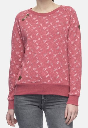 Žena ve světle růžovém svetru s dlouhým rukávem s drobným bílým květinovým vzorem a dekorativními hnědými knoflíky u límce a spodního lemu, v kombinaci se šedými džínami.