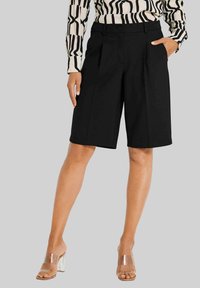 Schwarze maßgeschneiderte Shorts mit vorderen Falten, seitlichen Taschen und einem glatten Stofffinish, kombiniert mit klaren Absatzsandalen.