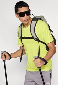 Φωτεινό κίτρινο κοντομάνικο πουκάμισο, γκρι σορτς, ανθεκτικό σακίδιο πλάτης, μαύρα μπαστούνια πεζοπορίας με γκρι λαβές και σκούρα γυαλιά ηλίου.