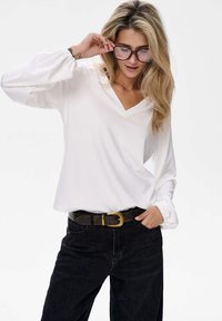 Weißes V-Ausschnitt-Oberteil mit langen Puffärmeln, kombiniert mit hochtaillierten schwarzen Jeans und einem braunen Gürtel mit einer goldenen Schnalle.