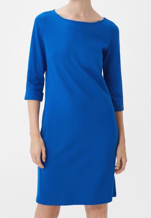 Kobieta ubrana w niebieską sukienkę do kolan z rękawami o długości do łokci i okrągłym dekoltem na jednolitym białym tle.