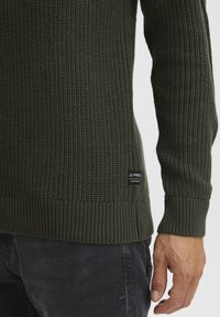 Dunkelgrüner, gerippter Strickpullover mit langen Ärmeln, der eine strukturierte Oberfläche aufweist und ein schwarzes Etikett auf der unteren linken Seite hat.