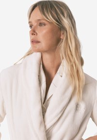 Biała, pluszowa szlafrok z kołnierzem typu szal, z teksturowaną powierzchnią i haftowanymi detalami. W połączeniu z jasnym, zapinanym na guziki koszulą.