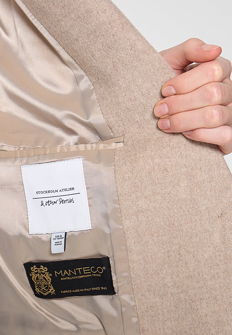 Beige Wollmantel-Innenfutter mit Satinfutter, zeigt Etiketten von 'Stockholm Atelier' und 'Manteco'. Strukturierter Stoff, hochwertige Ausführung.