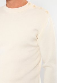 Kremowy sweter z dzianiny z okrągłym dekoltem, teksturowaną tkaniną i trzema guzikami na ramieniu.