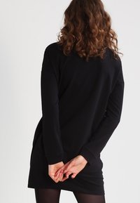 Czarna, długi rękaw sukienka o luźnym fasonie, z okrągłym dekoltem i zaokrąglonym dołem. Materiał wydaje się gładki i elastyczny.