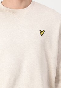 Beżowa bluza z okrągłym dekoltem, z małym żółtym emblematem ptaka wyhaftowanym na lewej piersi.
