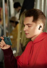 Wireless earbuds in a black color, worn by a person in a red hoodie, holding a green smartphone while seated in a transit setting.