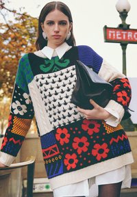 Pull en tricot surdimensionné avec des motifs variés en bleu, vert, noir, blanc, orange et rouge. Le mannequin tient un sac à main noir.