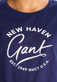 Námořnická modrá bavlněná tričko s bílým textem: "NEW HAVEN Gant EST. 1949 NHCT U.S.A." Kulatý výstřih a krátké rukávy.