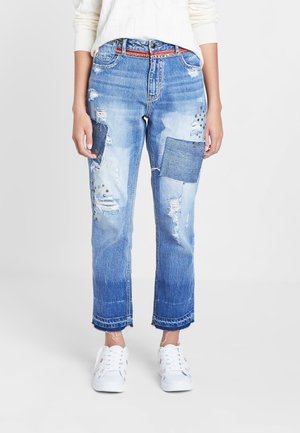 Niebieskie przetarte dżinsy z dużymi łatami i nitowanymi detalami, noszone z białymi trampkami i białym swetrem.