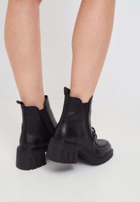 Botas de piel negras hasta el tobillo con suela gruesa, cremallera lateral, punta redondeada y tirador en la parte trasera. Textura suave y detalles de diseño simples.