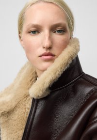 Giacca in pelle marrone con colletto in pelliccia beige. Presenta una texture liscia e una chiusura con zip, evidenziando un design moderno e caldo.