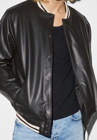 Veste aviateur en cuir noir avec boutons-pression, col côtelé et ourlet à rayures. Portée sur un haut noir et associée à un jean bleu clair.