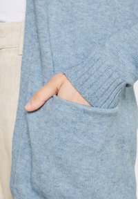 Main insérée dans la poche d'un pull en tricot bleu clair, pantalon beige partiellement visible en arrière-plan.