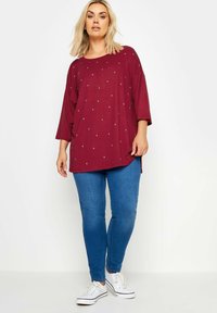 Burgundy oversized tunika med korta ärmar, prydd med silverstjärnor; matchad med blå skinny jeans och vita sneakers.