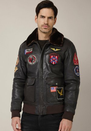 Brązowa skórzana kurtka bomberka z futrzanym kołnierzem, posiadająca wiele haftowanych naszywek oraz dwie przednie kieszenie; ściągacze przy mankietach i u dołu.