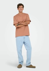 Lichtbruin katoenen t-shirt met korte mouwen, gecombineerd met lichtblauwe denim jeans. Model staat met over elkaar geslagen armen, draagt een paar bruine instap schoenen.