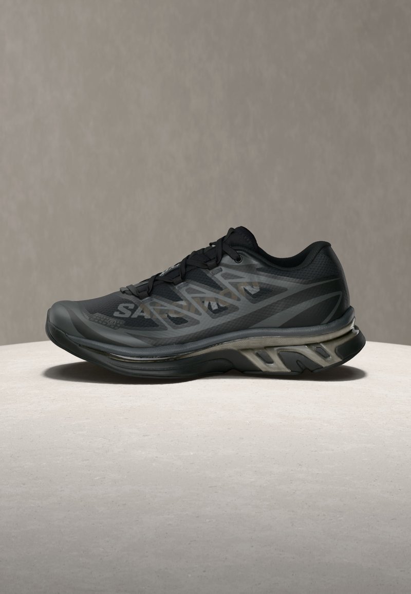 Black athletic shoe with geometric gray patterns, thick cushioned sole, and laces, shown from the side on a light surface against a neutral background.