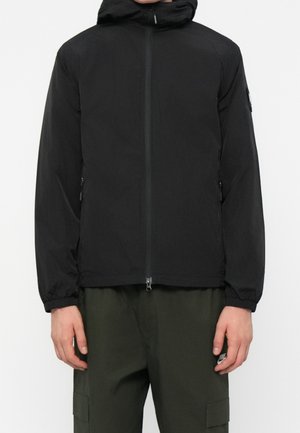 Homme portant une veste noire à capuche avec fermeture éclair devant et un pantalon cargo vert, debout devant un fond blanc.