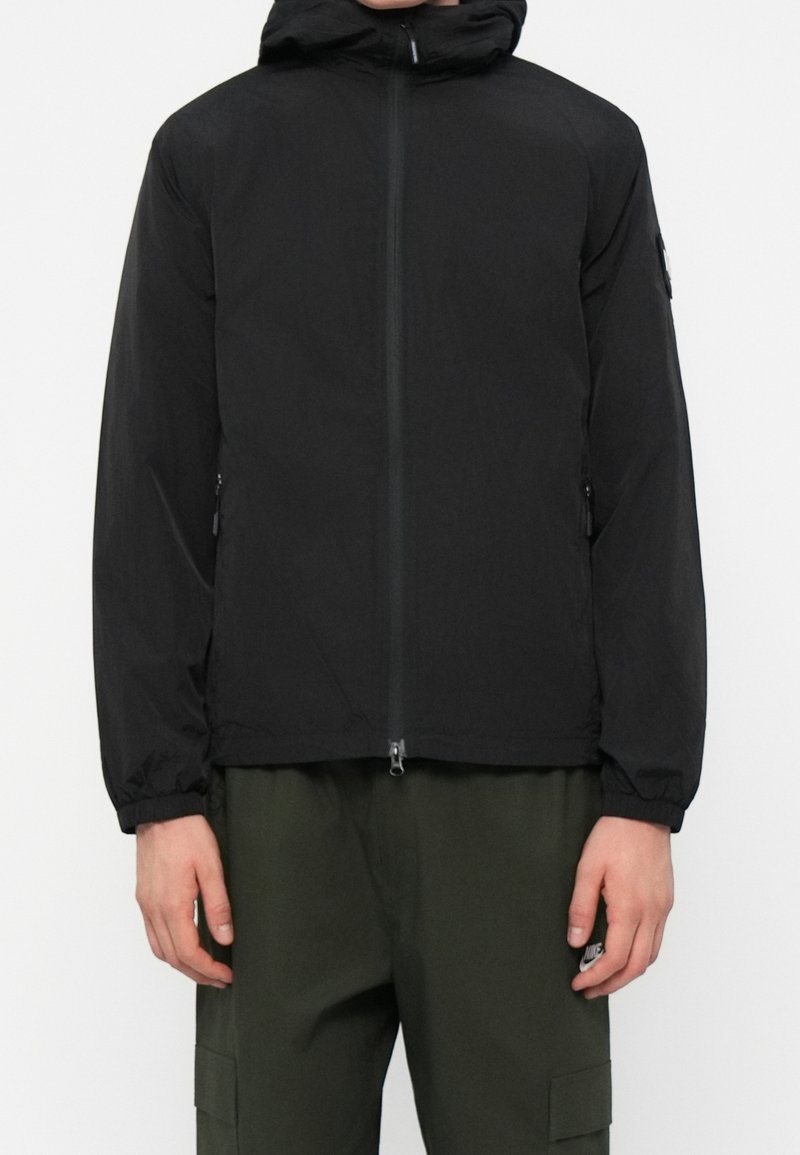 Homme portant une veste noire à capuche avec fermeture éclair devant et un pantalon cargo vert, debout devant un fond blanc.