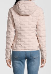 Veste matelassée rose clair avec capuche, présentant une coupe ajustée et des poignets élastiques, associée à un jean en denim bleu. Logo visible sur l'ourlet.