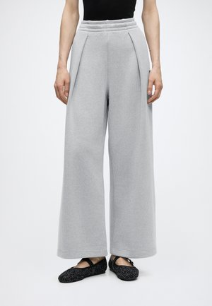 Personne portant un pantalon gris clair taille haute à jambes larges avec plis, associé à des ballerines noires à brides, debout devant un fond clair uni.