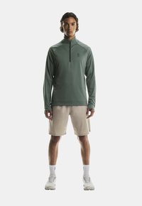 Zaļš garām piedurknēm sporta tops ar puscirtni, kombinēts ar gaiši bežām šortiem. Izkārtots ar logotipu uz krūtīm, gludu tekstūru un minimālu dizainu.